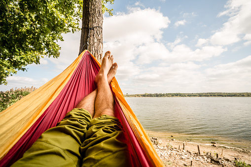 Relaxing in the hammock at the beach under a tree, summer day. Barefoot man laying in hammock, looking on a lake, inspiring landscape