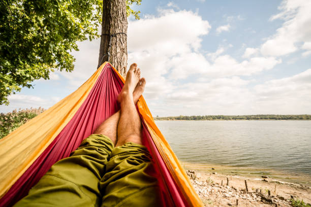 Relaxing in the hammock at the beach under trees, summer day Relaxing in the hammock at the beach under a tree, summer day. Barefoot man laying in hammock, looking on a lake, inspiring landscape hammock stock pictures, royalty-free photos & images