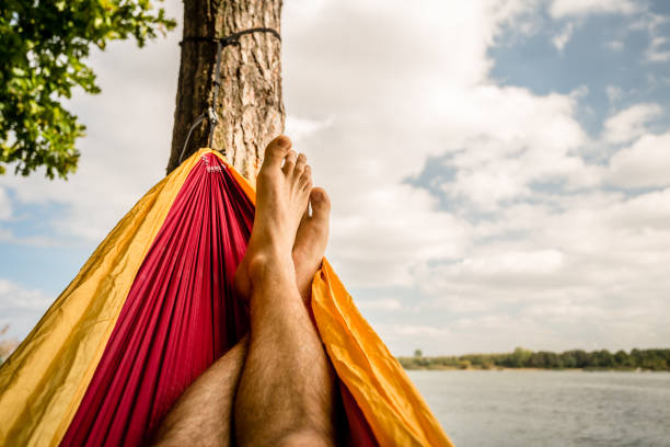 Relaxing in the hammock at the beach under a tree, summer day. Barefoot man laying in hammock, looking on a lake, inspiring landscape
