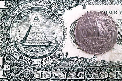 Close-up on a 1 dollar bill with the focus on the Eye Of Providence and on the eagle of 25 cents coin.