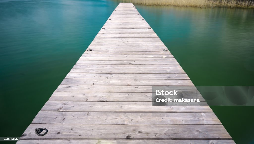 wooden pier jutting out into a turquoise and blue lake with golden reeds in the background A wooden pier jutting out into a turquoise and blue lake with golden reeds in the background Anchored Stock Photo
