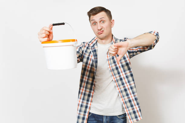 istock Young dissatisfied handsome man holding empty paint bucket with copy space and showing thumb down isolated on white background. Instruments, tools for renovation apartment room. Repair home concept. 960547724