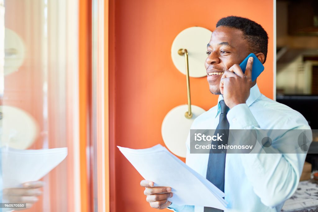 Working with client by phone Happy young agent in formalwear talking by smartphone while looking through window of his office Using Phone Stock Photo