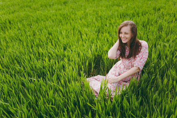 istock Young brunette smiling beautiful woman in light patterned dress sitting on grass looking aside keeping hand near hair in sunny weather in field on bright green background. Lifestyle, leisure concept. 960574666