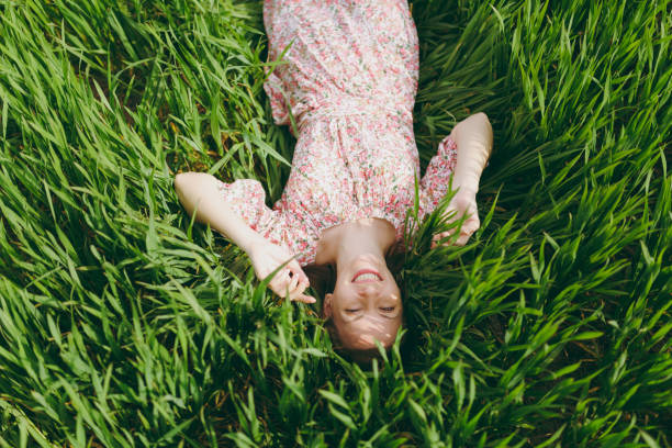 istock Young smiling pretty woman in light patterned dress lying on grass spreading hands looking up resting in sunny weather in field on bright green background. Spring nature. Lifestyle, leisure concept. 960574890