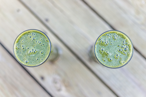 Macro closeup of two green vegetable fruit smoothies pair couple in glasses outside outdoors on wooden deck background for healthy raw vegan breakfast