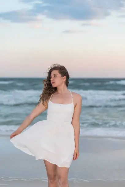 Young woman in white dress on beach sunset in Florida panhandle with wind, ocean waves, side profile looking away