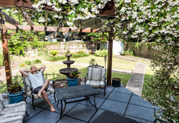 młody człowiek leżący na patio leżak w odkrytym ogrodzie kwiatowym wiosną w podwórku ganek domu szczęśliwy uśmiecha się w zen z fontanną, pergola baldachim altanka, stół, rośliny, sofa - fountain in garden zdjęcia i obrazy z banku zdjęć