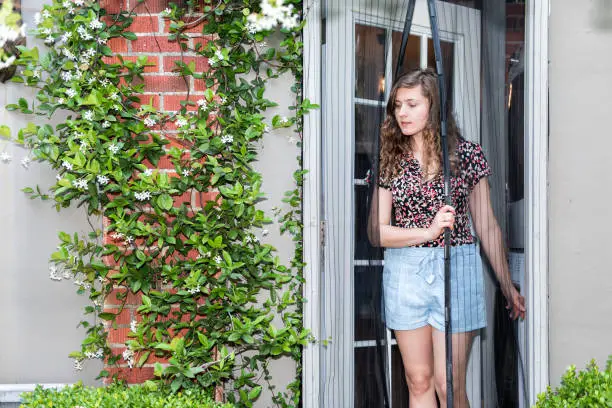 Young woman exiting house entering outdoor spring garden with hanging plant flowers covering brick wall in backyard porch of home by door, insect net