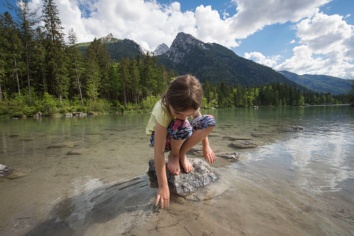A girl with a yellow shirt stands in a mountain lake and enjoys playing. In the background stands the hochkalter mountain
