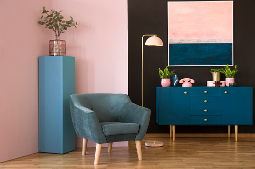 Blue suede armchair against pink wall in living room interior with cabinet under painting