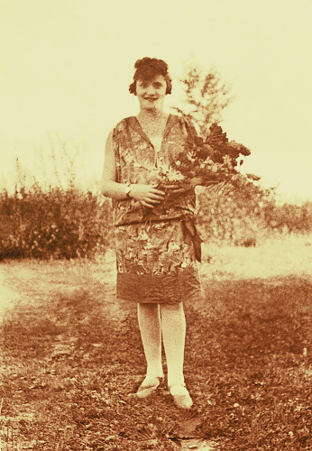 Vintage sepia toned photo of a young woman from the beginning of the twentieth century standing outdoors in the nature with flowersin her hands.
