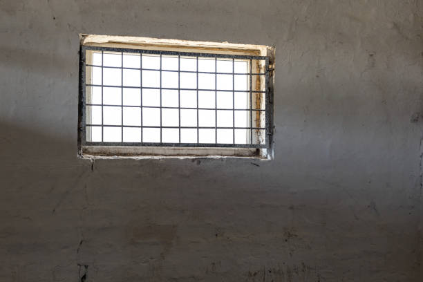 istock Old white wall with window covered by iron grate. Sun shining  through grill. Prison room interior . Enlightment concept. Copyspace 960637014