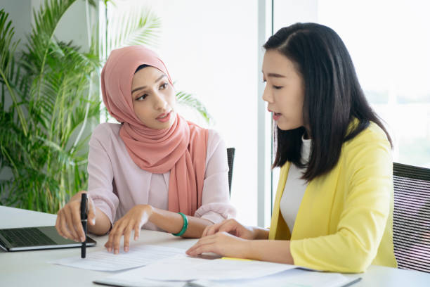 Malaysian woman explaining document Two young businesswomen in office looking at paperwork, Malay woman holding men and looking towards Chinese co-worker malaysia office workers stock pictures, royalty-free photos & images