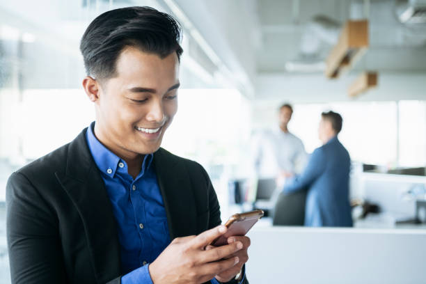Young businessman texting on smartphone at work Malaysian man in his 20s looking down and smiling as he uses his mobile phone in modern office rockabilly hair men stock pictures, royalty-free photos & images