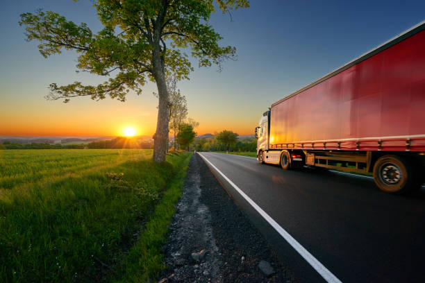 camion conduite sur la route goudronnée entre les arbres dans un paysage rural au coucher du soleil - semi truck truck highway red photos et images de collection