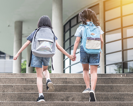 Back to school education concept with girl kids (elementary students) carrying backpacks going to class holding hand in hand together walking up school building stair happily