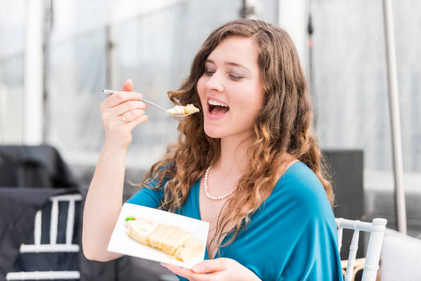 elegante mujer sonriente de joven feliz comiendo pastel, abrir la boca con un tenedor durante la cena de postre boda recepción, sentado en la mesa - women spoon tasting elegance fotografías e imágenes de stock