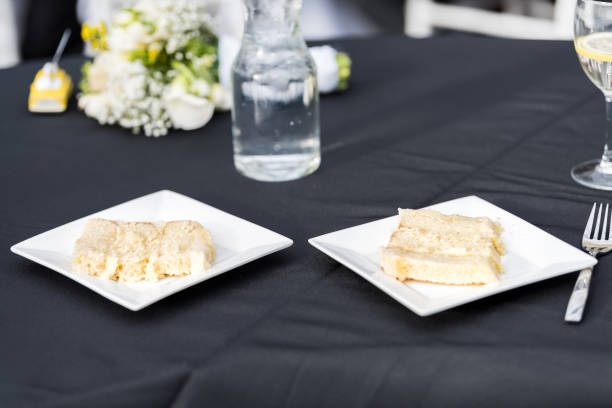Glass with lemon slice and water in restaurant or wedding reception black table tablecloth, two plates with cake slices, bouquet flowers, forks
