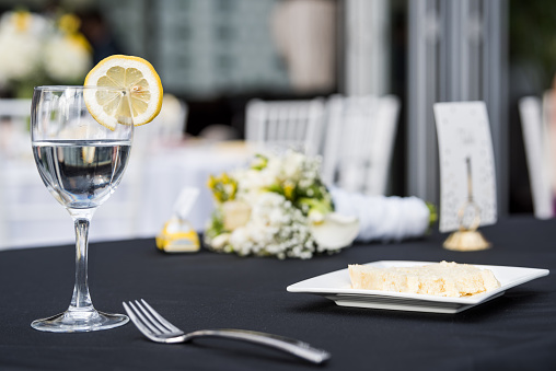 Closeup of one glass with lemon slice and water in restaurant or wedding reception black table tablecloth, plate with cake slice, bouquet flowers, fork, chairs, dishes
