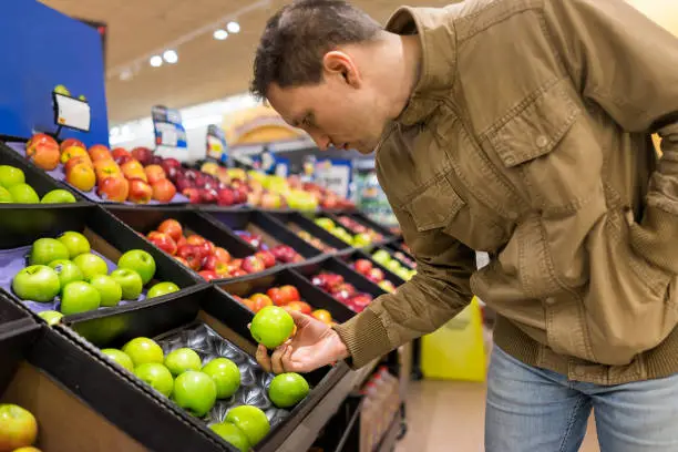 Many varieties assorted apples on display shelf in grocery store boxes in aisle, supermarket inside, man person customer holding choosing granny smith green fruit