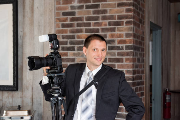 un jeune heureux souriant photographe de mariage en costume-cravate debout avec appareil photo, flash externe, trépied à l’intérieur du bâtiment restaurant salle intérieure - photographer camera tripod paparazzi photographer photos et images de collection