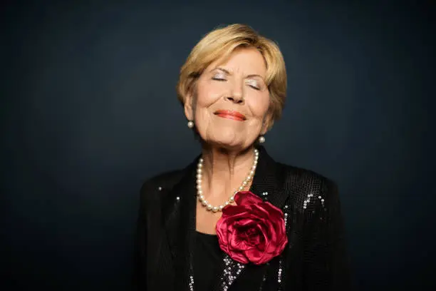 Smiling senior woman with eyes closed. Fashionable elderly female is wearing pearl jewelry and artificial pink rose. She is against colored background.