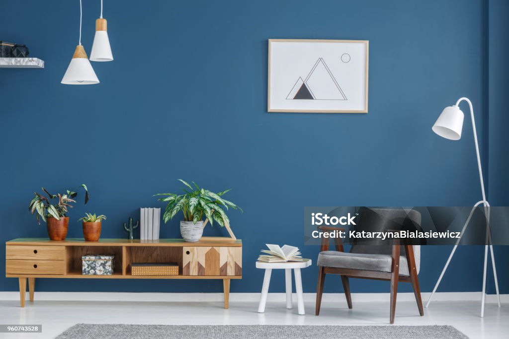 Small wooden cupboard Small wooden cupboard with books, fresh plants and decor in blue living room interior with grey armchair and simple poster Living Room Stock Photo