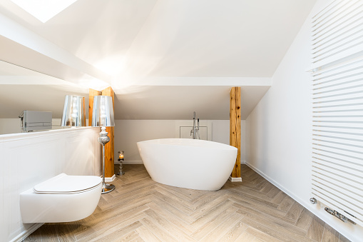 Modern white attic bathroom interior with oval ceramic bathtub, wall heater and exposed wooden beams