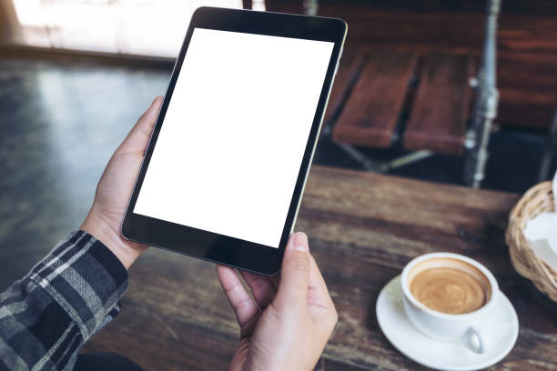 istock Mockup image of hands holding black tablet pc with white blank screen and coffee cup on table background 960772876