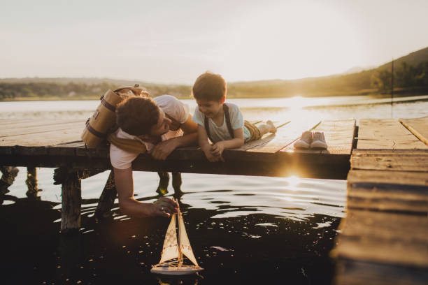 Sailing boat with my dad Photo of a young man and his son sailing the toy boat into the lake water while sitting on the dock. toy boat stock pictures, royalty-free photos & images