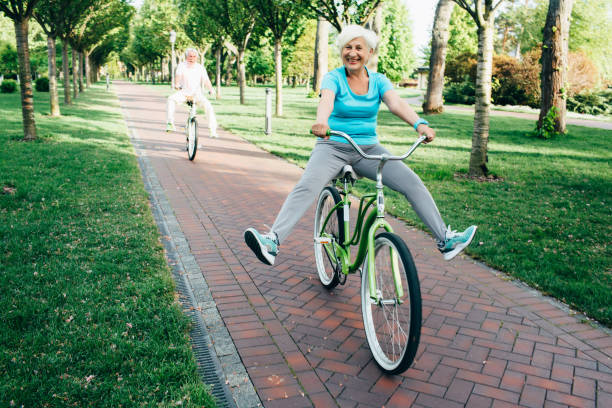 elderly woman having fun cycling happy senior couple riding their bikes on the park, funny lifting their legs up young at heart stock pictures, royalty-free photos & images