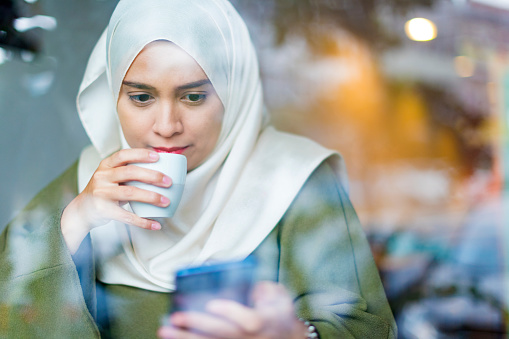 Young woman in a hijab looking over her messages at a cafe while sipping some tea.