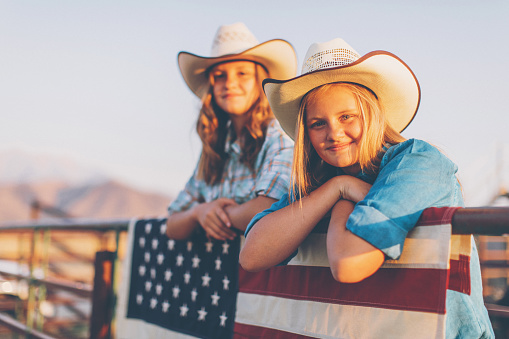 Young teenage cowgirls and sisters in cowboy hats are leaning on a metal fence. An American flag is hanging from the fence. They are smiling and looking at the camera. They love celebrating the Fourth of July. Image taken in Utah, USA.
