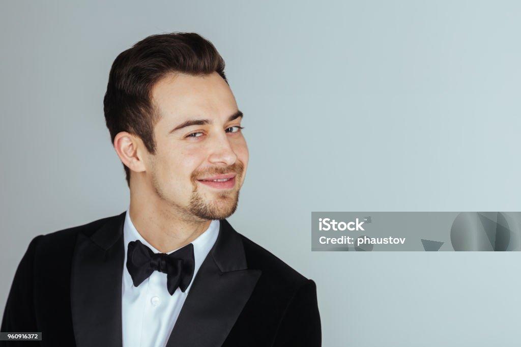 Young handsome man in a tuxedo,  looking at the camera Portrait of a young handsome man in a tuxedo,  smiling and looking at the camera, against plain studio background. Mischief Stock Photo