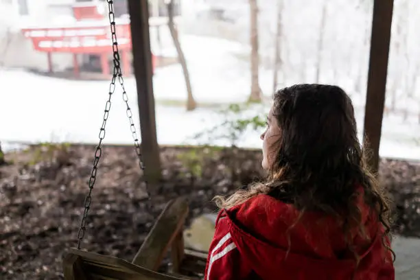 Closeup of young woman sitting on swing under wooden deck of house on backyard in neighborhood with snow during blizzard white storm, snowflakes falling in Virginia suburb, single family home
