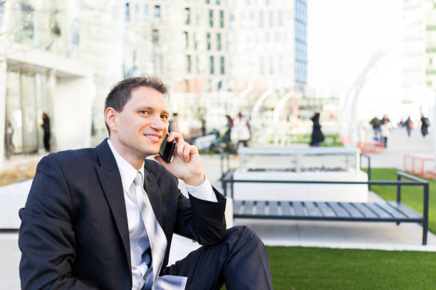 istock Handsome, attractive young businessman sitting on bench, using talking holding smartphone phone mobile cellphone smiling in suit and tie, cheerful on interview break in urban park 960927794