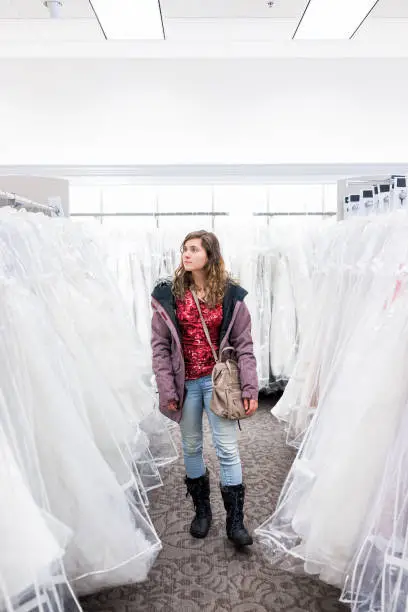 Young woman looking at wedding dress in boutique discount store, many white garments hanging on rack hangers row