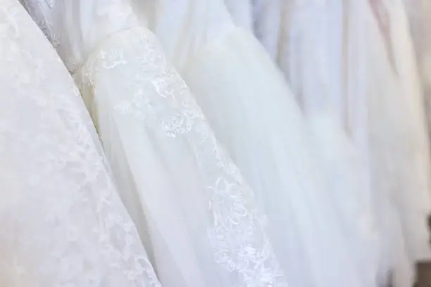 Many wedding dresses in boutique discount store, white garments hanging on rack hangers row closeup with white lace, tulle, design