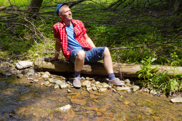 istock Adult farmer sitting and resting on tree trunk next to a flowing stream 961012656