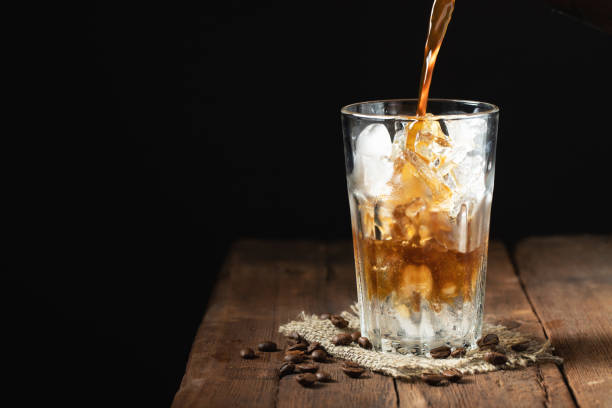 istock Ice coffee in a tall glass over and coffee beans on a old rustic wooden table. Cold summer drink on a dark background with copy space. The process of pouring drink from a coffee pot into a glass 961020972