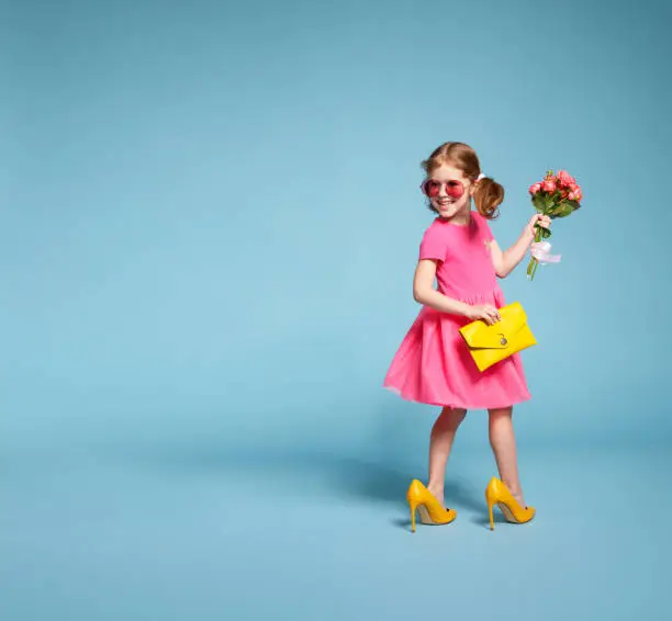 Photo of funny child girl fashionista in big mother's   shoes on colored background