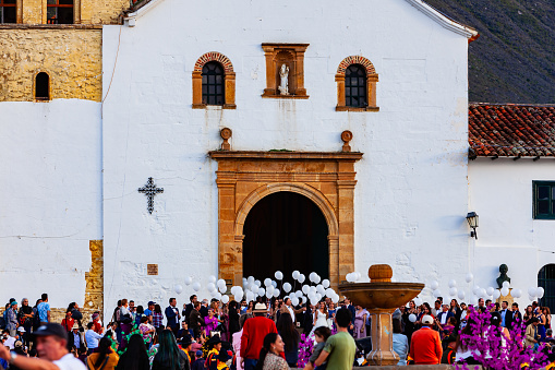 Villa de Leyva, Colombia - November 25, 2017: A wedding has just been concluded in the church on the main plaza in the historic town of Villa de Leyva. White balloons are about to be released in celebration. Founded in 1572, and located at just over 7000 feet above mean sea level on the Andes Mountains, the 16th Century town of Villa de Leyva was declared a National Monument in 1954 to protect its colonial architecture and heritage. It is located in the Department of Boyaca, in the South American country of Colombia. Seen in the image are local residents and some Colombian tourists visiting the town, all curious witnesses to the wedding. In the foreground is the stone fountain on the centre of the square. Photo shot in the late afternoon sunlight; horizontal format. Copy space. Camera: Canon EOS 5D MII. Lens: Canon EF 24-70 F2.8L USM. Canon EF 70-200mm F2.8L IS USM.