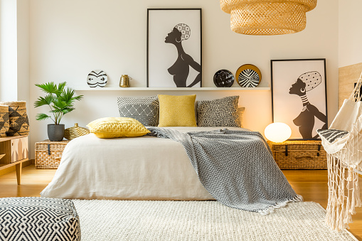 Yellow and patterned pillows on bed in modern warm bedroom interior with posters and plant