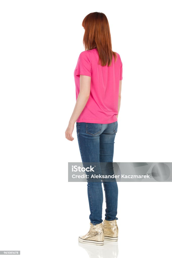 Rear Side View Of Young Standing Woman In Pink Shirt Young woman in pink t-shirt, jeans and gold sneakers is standing and looking away. Rear side view. Full length studio shot isolated on white. Rear View Stock Photo