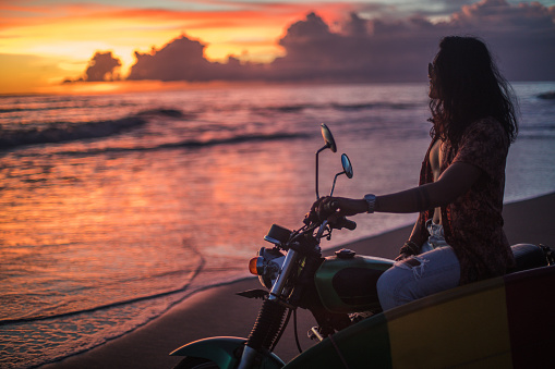 One man, handsome hippie surfer sitting on motorcycle on the beach in sunset.