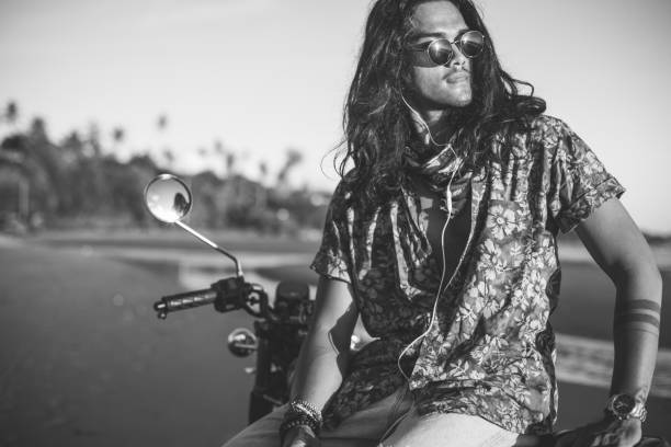 One man, handsome hippie biker sitting on motorcycle on the beach in sunset, listening music on headphones, black and white.