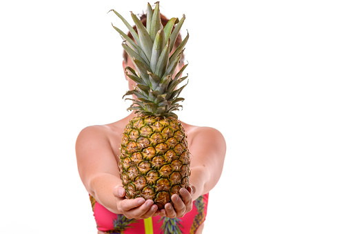 Upper body portrait of woman in swimsuit holding pineapple in front of face, white background.