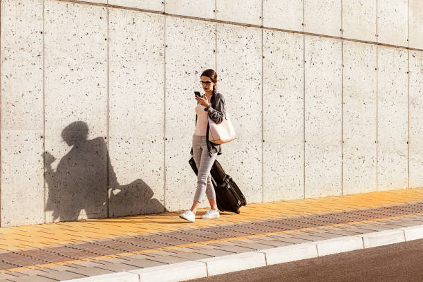 istock Young woman using her smart phone walking beside the concrete wall and pulling a small wheeled luggage with a briefcase on it 961271570