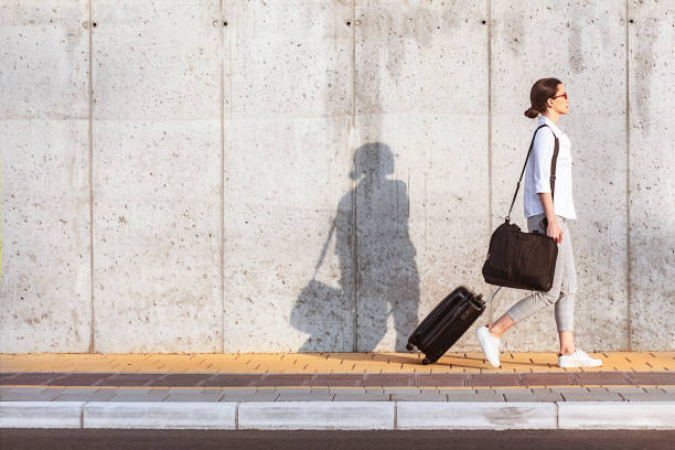 Young woman walking on a sidewalk beside the concrete wall and pulling a small wheeled luggage